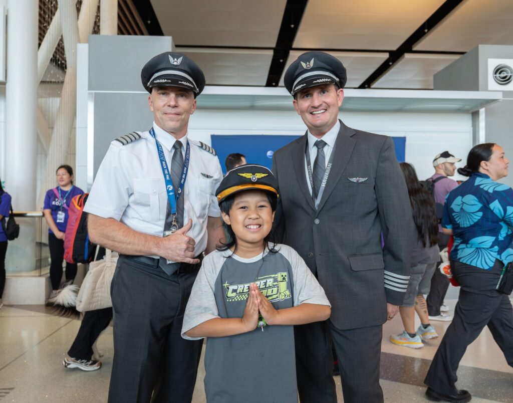 Engelli prova uçuşuna katılan bir çocuk pilot şapkasıyla iki Hawaiian Airlines pilotunun arasında gülümsüyor