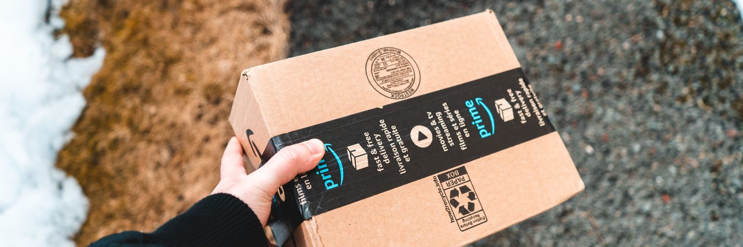 Karlı bir yolda siyah montlu birinin tuttuğu Amazon Prime karton kolisi.