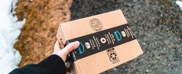Karlı bir yolda siyah montlu birinin tuttuğu Amazon Prime karton kolisi.