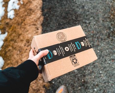 Karlı bir yolda siyah montlu birinin tuttuğu Amazon Prime karton kolisi.