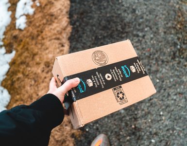Karlı bir yolda siyah montlu birinin tuttuğu Amazon Prime karton kolisi.